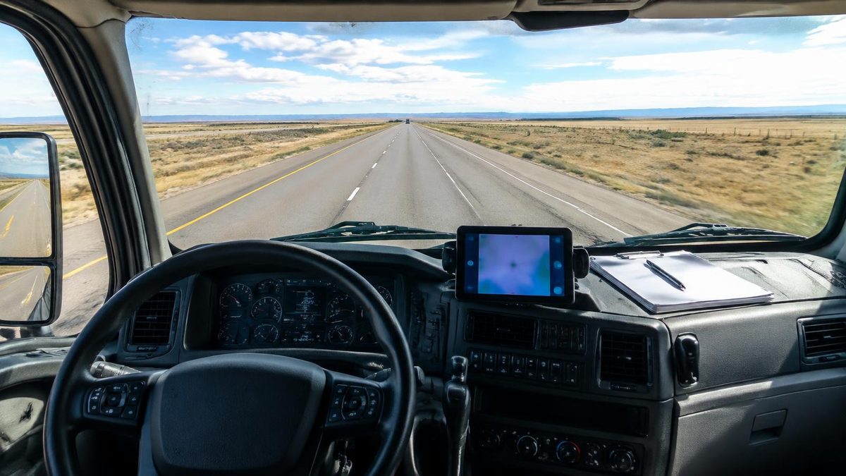 View from an unbranded truck cab over a tablet and paperwork toward an open plains highway